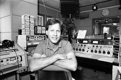 Rick Dees sitting with arms crossed inside the KIIS-FM studio surrounded by broadcast equipment