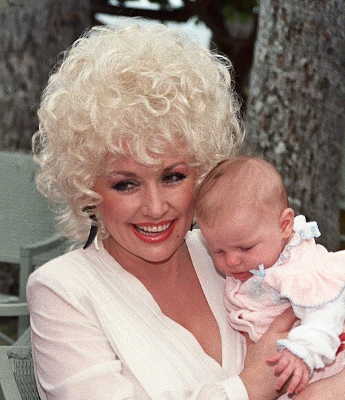 Dolly Parton smiling while holding a baby outdoors