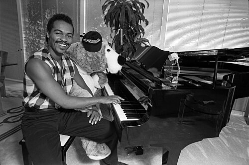 Ray Parker Jr. seated at a grand piano with a stuffed toy monkey wearing a Ghostbusters cap, 1984