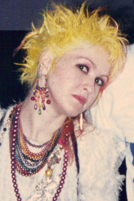 A close up portrait of Cyndi Lauper with spiky yellow hair and layered beaded necklaces
