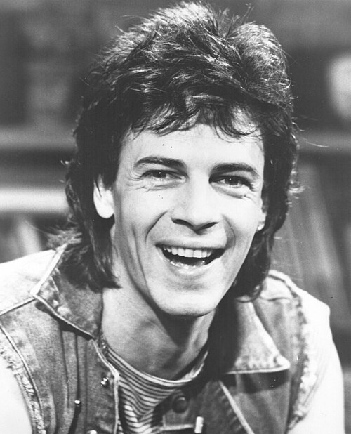A smiling black and white portrait of Rick Springfield with tousled hair, wearing a denim vest