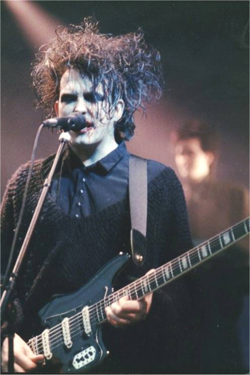 Robert Smith of The Cure singing into a microphone and playing guitar on stage, 1989