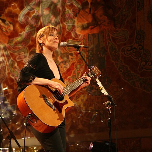 Suzanne Vega singing into a microphone while playing acoustic guitar on stage