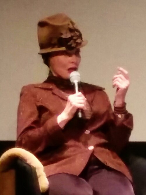 Toni Basil speaking into a microphone at an event, wearing a wide brimmed hat