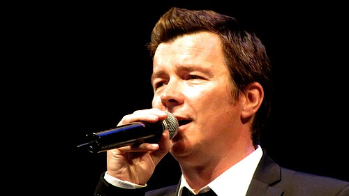 Rick Astley singing into a microphone on stage