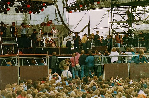 UB40 performing on an outdoor festival stage in front of a large crowd, 1987