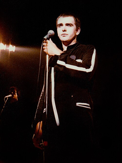 Peter Gabriel singing into a microphone on stage, wearing a black and white jacket