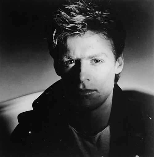 A moody black and white portrait of Bryan Adams from 1984, wearing a leather jacket