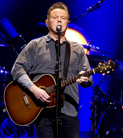 Don Henley singing into a microphone while playing acoustic guitar on stage
