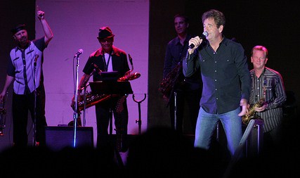 Huey Lewis singing at the microphone on stage, with the News performing behind him