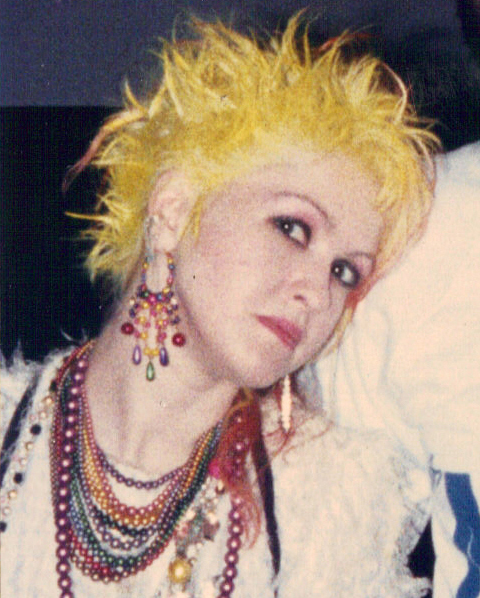 A close up portrait of Cyndi Lauper with spiky yellow hair and layered beaded necklaces, 1985